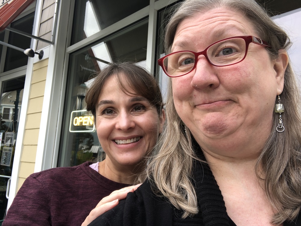 Sue with a goofy expression and a friend smiling normally in front of a coffee shop.
